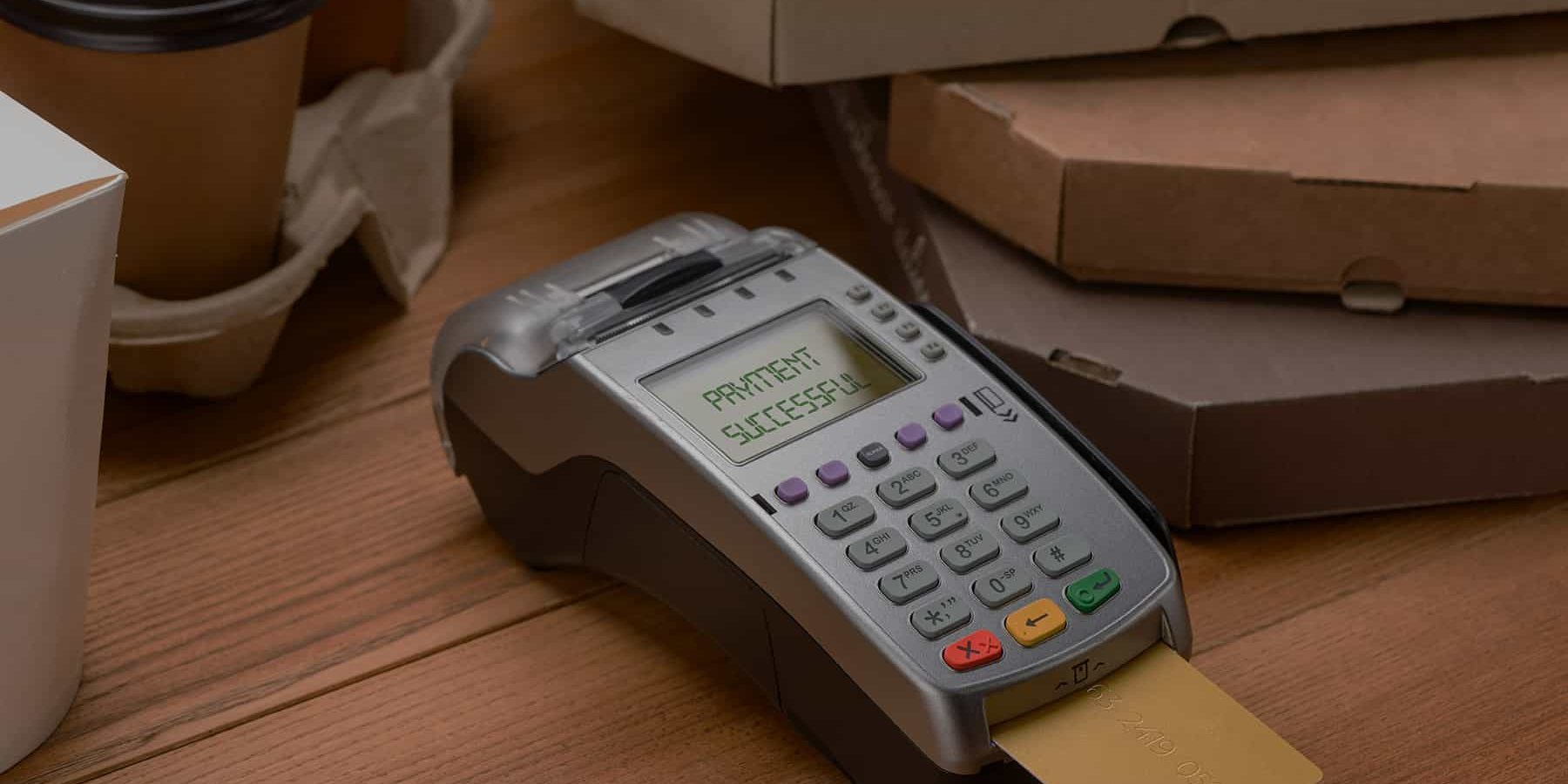 Paying for coffee and pizza. POS terminal with inserted credit card beside take-away cups and boxes.